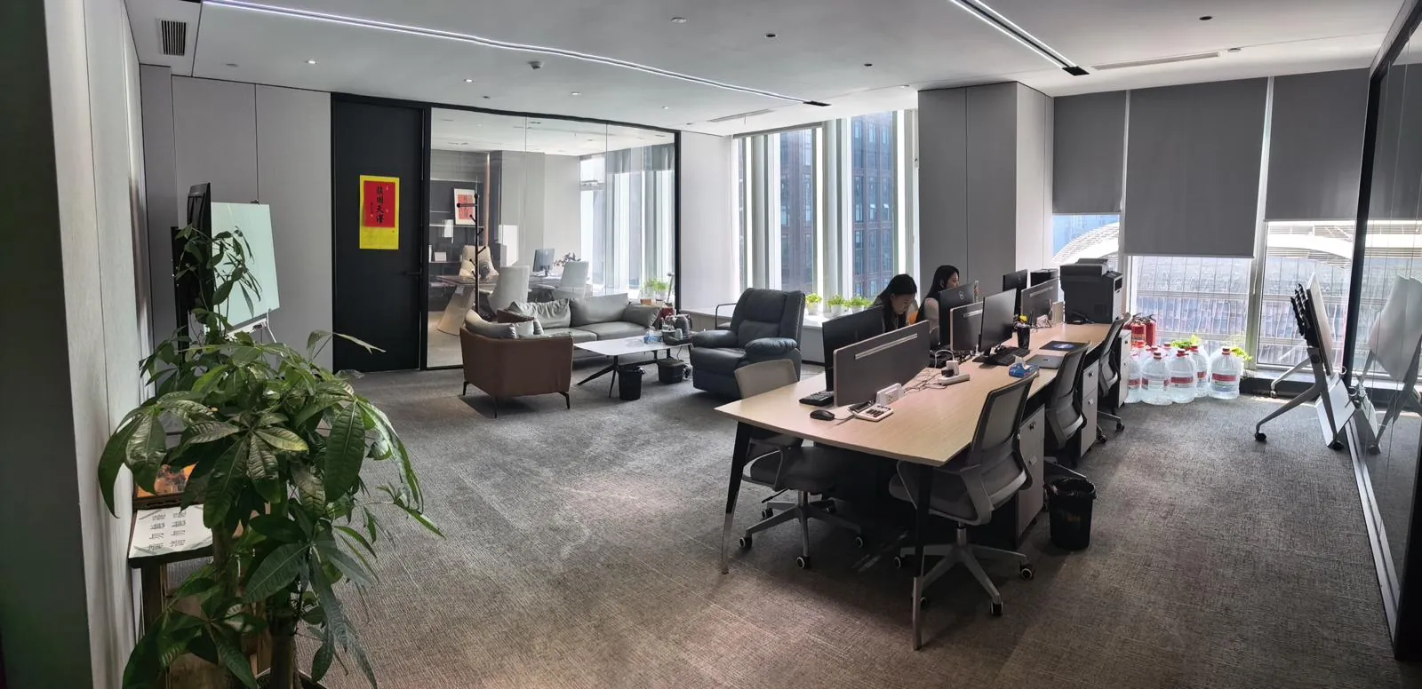 Open-plan office with staff at their desks and a lounge beside tall windows